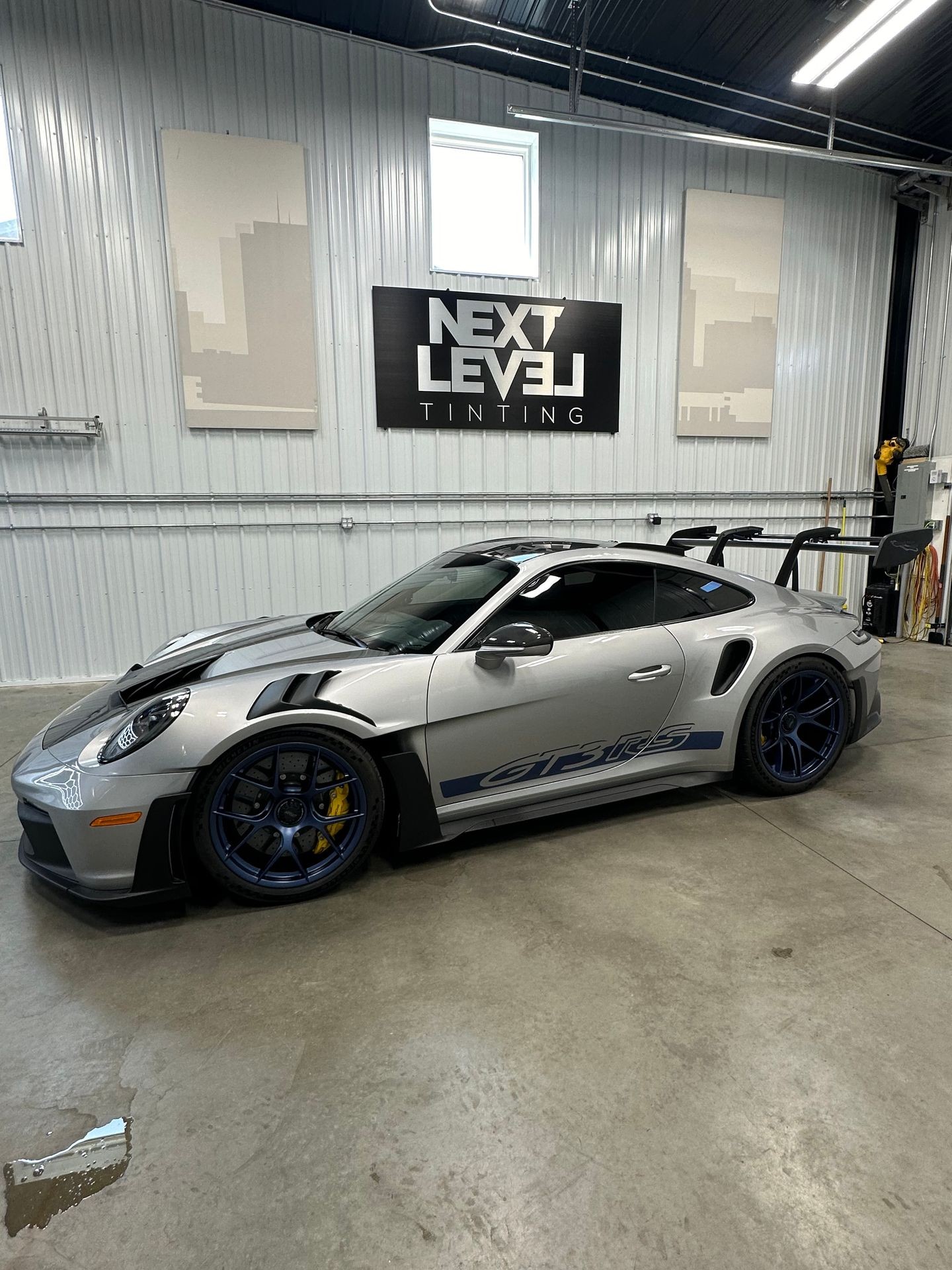 Silver sports car parked in a garage with 'Next Level Tinting' sign on the wall.